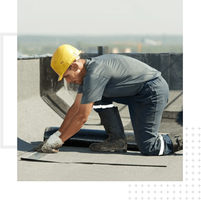 A man wearing a hard hat and gloves using a roll of roofing material.