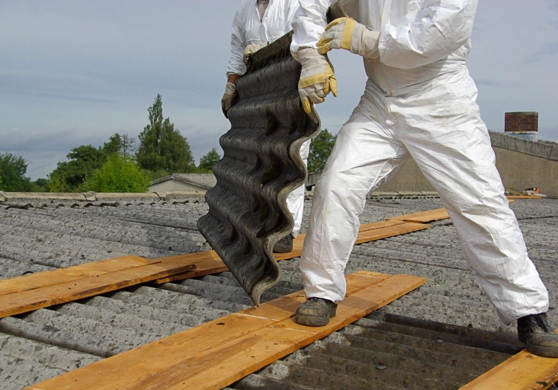 A man in white protective gear carrying a black piece of roofing material.